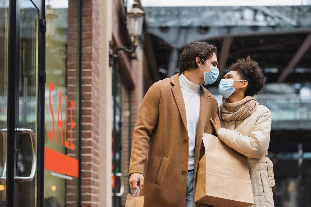 pareja interracial joven en máscaras médicas mirándose y caminando con bolsas de compras en el centro comercial  - Foto, imagen