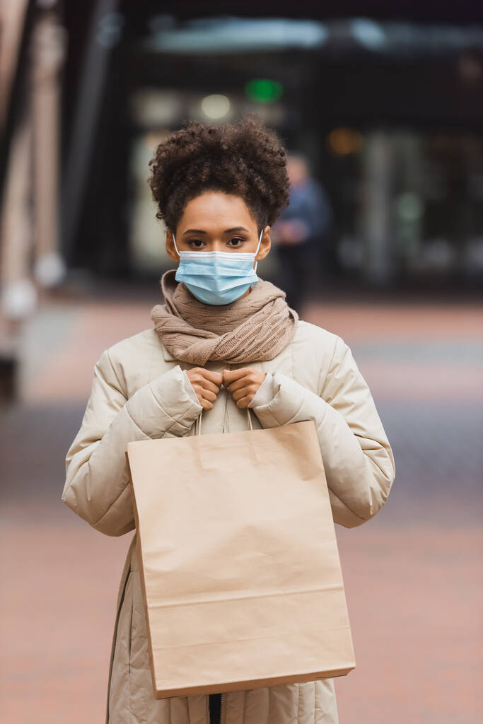 brunette afro-américaine en masque médical tenant sac à provisions dans le centre commercial  - Photo, image