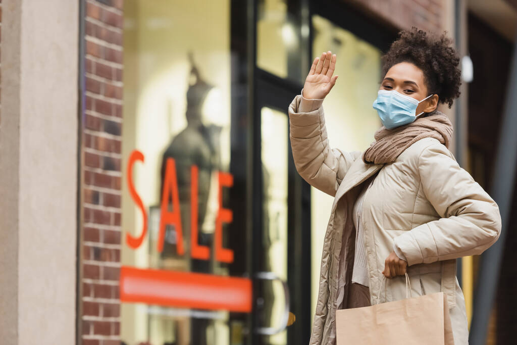 brünette afrikanisch-amerikanische Frau in medizinischer Maske mit Einkaufstasche und winkender Hand in Einkaufszentrum  - Foto, Bild