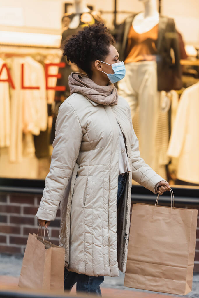 μελαχρινή Αφροαμερικανή γυναίκα με ιατρική μάσκα περπατώντας με τσάντες για ψώνια στο εμπορικό κέντρο  - Φωτογραφία, εικόνα