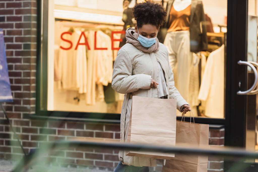 joven mujer afroamericana en máscara médica sosteniendo bolsas de compras en el centro comercial  - Foto, imagen