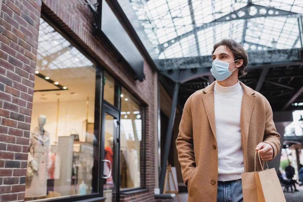 giovane in maschera medica e cappotto beige che cammina con shopping bag nel centro commerciale - Foto, immagini