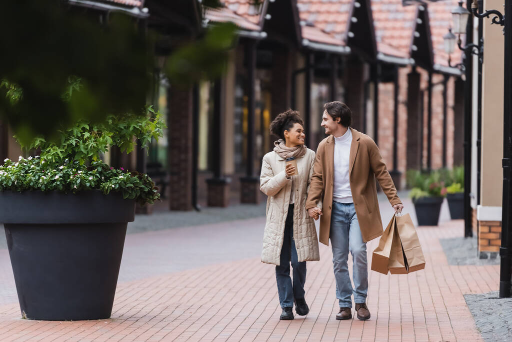feliz casal multiétnico em casacos de mãos dadas enquanto caminhava com compras perto do shopping - Foto, Imagem