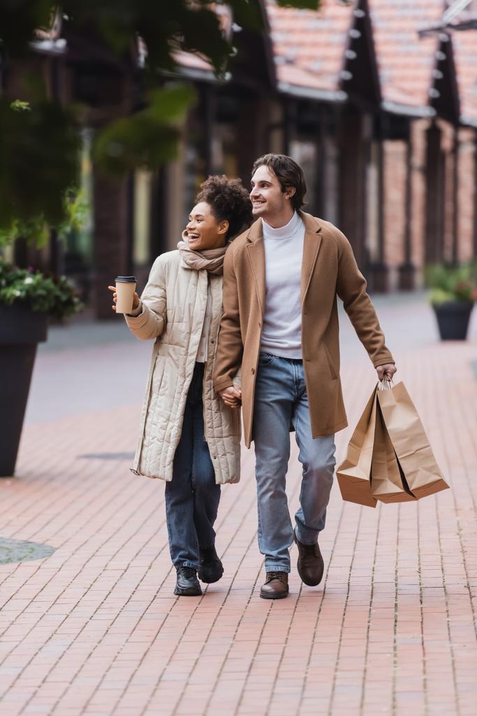 cheerful multiethnic couple in coats holding hands while walking with purchases near mall - Photo, Image