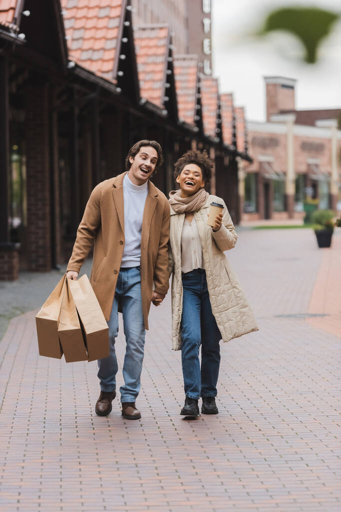izgatott multietnikus pár kabátban kéz a kézben sétálva vásárlás a bevásárlóközpont közelében - Fotó, kép