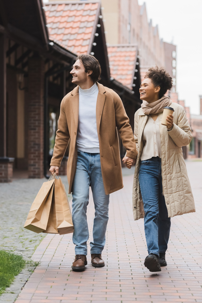 joyeux couple multiethnique en manteaux tenant la main tout en marchant avec des achats près du centre commercial - Photo, image