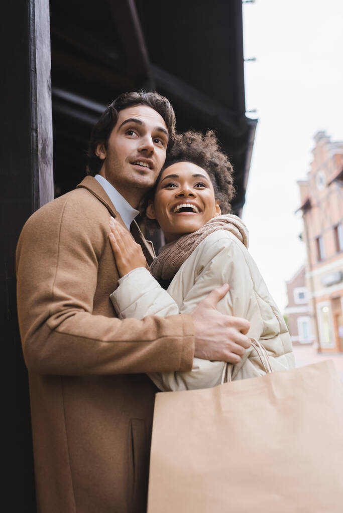 baixo ângulo vista de animado afro-americano mulher abraçando namorado com sacos de compras - Foto, Imagem