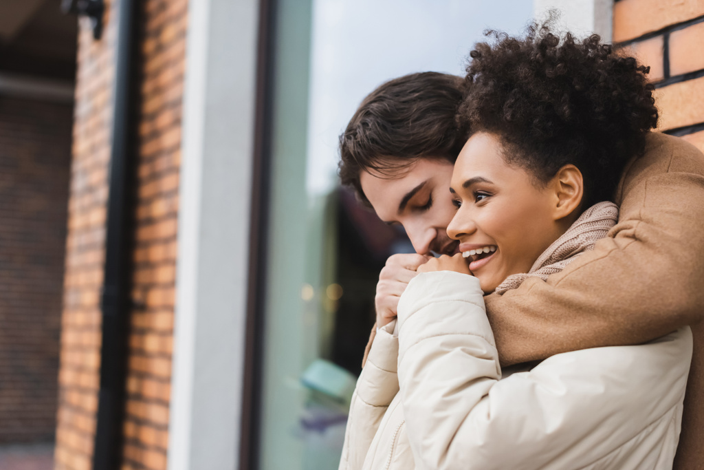 young man embracing and warming up cheerful african american girlfriend near building outdoors  - Photo, Image