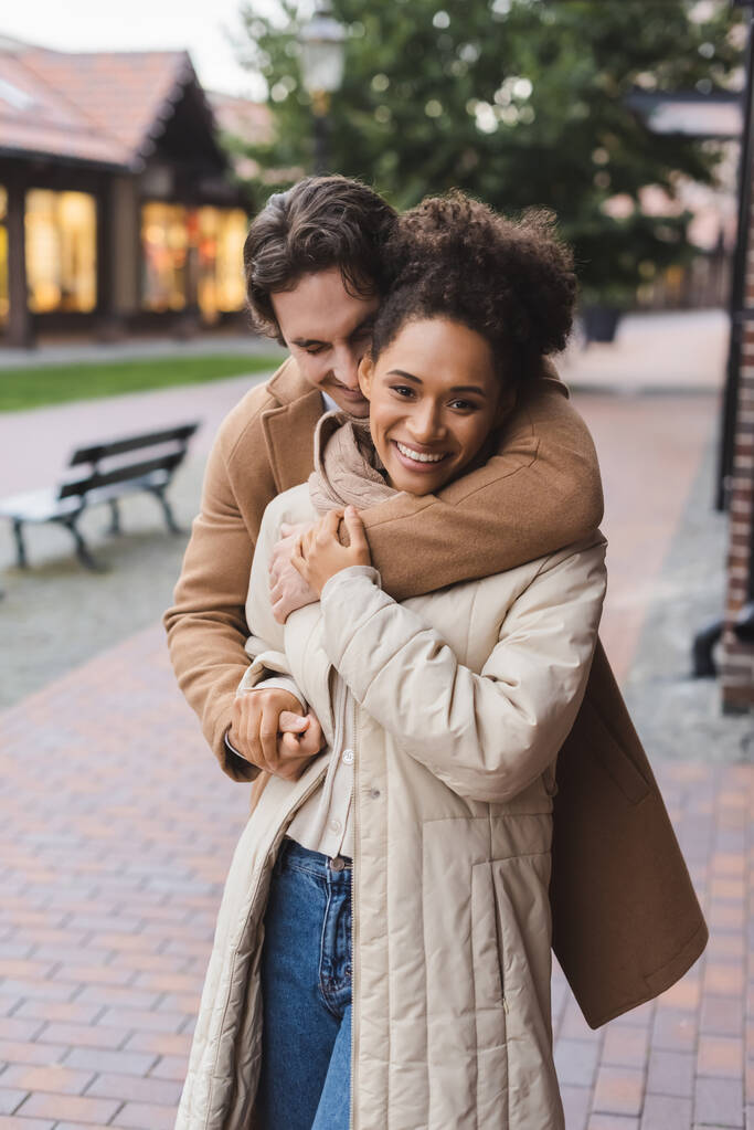 uomo sorridente abbracciando felice ragazza afro-americana vicino edificio all'aperto  - Foto, immagini