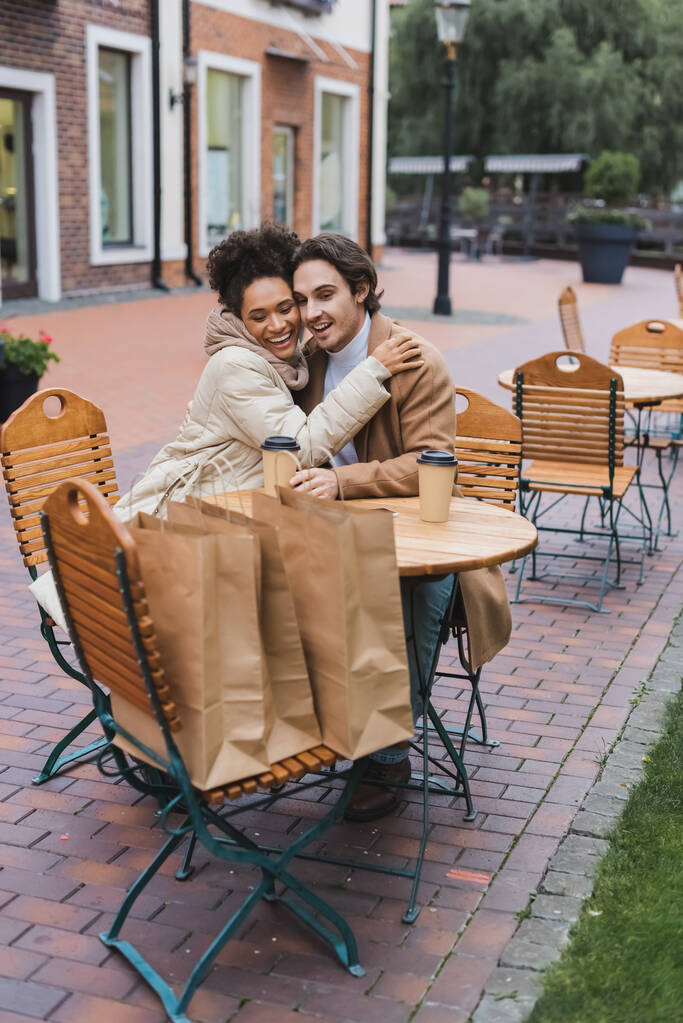 onnellinen rotujenvälinen pari syleilee lähellä paperi kupit ja ostoskassit ulkona kahvilassa  - Valokuva, kuva