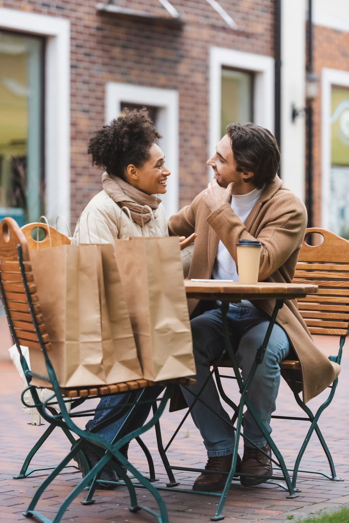 весела міжрасова пара розмовляє і дивиться один на одного біля паперової чашки і сумки для покупок у відкритій терасі кафе
  - Фото, зображення