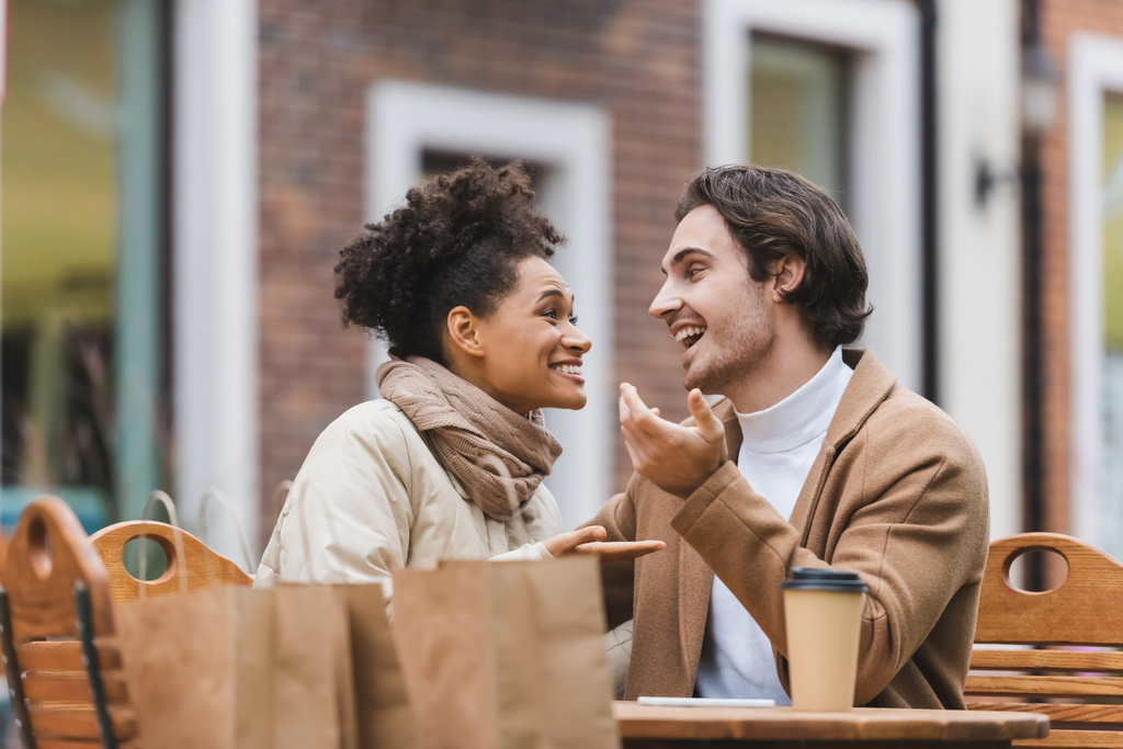alegre pareja interracial hablando y mirándose cerca de la taza de papel y bolsas de compras en la terraza de la cafetería al aire libre  - Foto, imagen