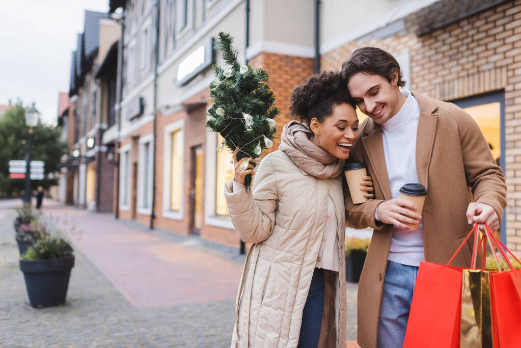 casal interracial espantado com café para ir, sacos de compras de Natal e pinho pequeno perto do shopping  - Foto, Imagem