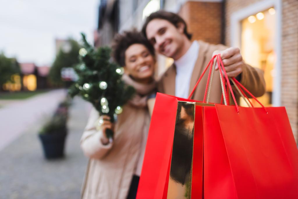 borrosa pareja interracial con bolsas de compras de Navidad cerca del centro comercial  - Foto, imagen
