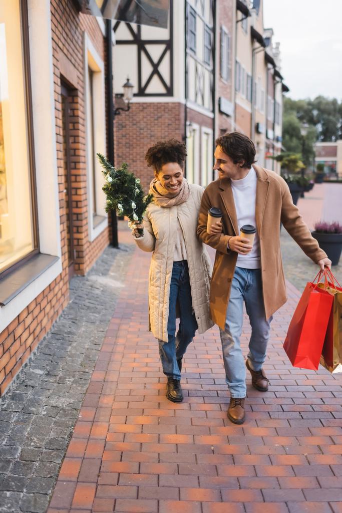 ευτυχισμένο πολυεθνικό ζευγάρι με καφέ για να πάει, τσάντες για ψώνια Χριστουγέννων και πεύκο περπάτημα κοντά στο εμπορικό κέντρο  - Φωτογραφία, εικόνα