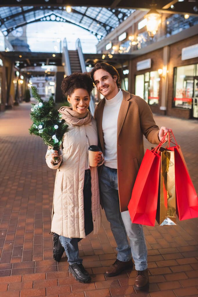feliz pareja multiétnica sosteniendo café para llevar, bolsas de compras de Navidad y pino en la noche - Foto, imagen