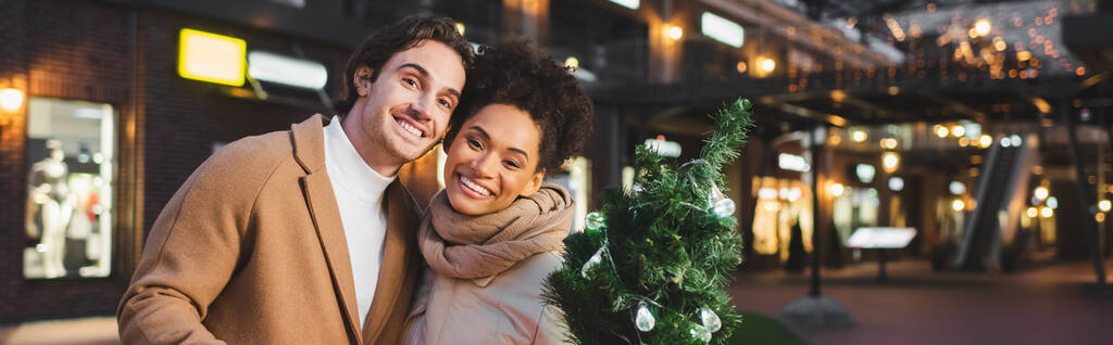 glückliches multiethnisches Paar mit kleiner Weihnachtskiefer am Abend, Banner - Foto, Bild