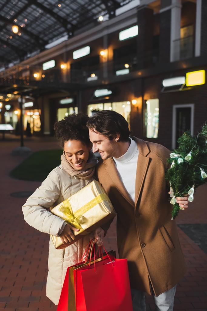 felice coppia multietnica tenendo borse della spesa di Natale, confezione regalo e pino vicino al centro commerciale in serata - Foto, immagini