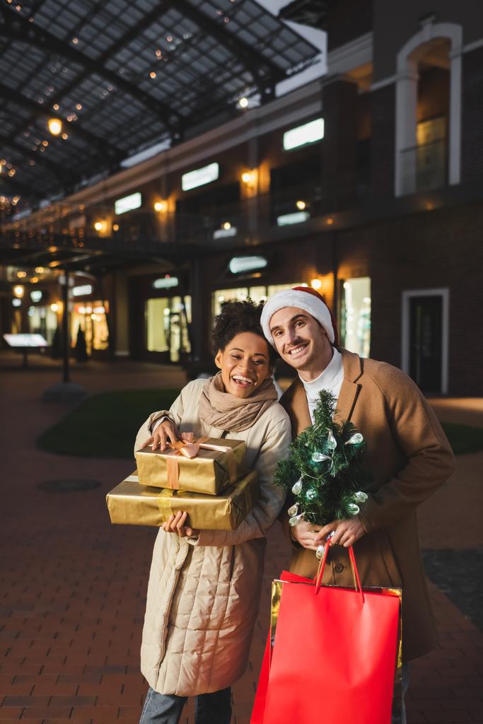boldog férfi télapó kalap gazdaság karácsonyi bevásárló táskák és fenyő közelében afro-amerikai barátnő ajándékokkal  - Fotó, kép