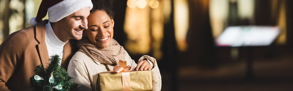 hombre feliz en sombrero de santa celebración de Navidad pequeño pino cerca de la novia afroamericana con caja de regalo, pancarta - Foto, Imagen