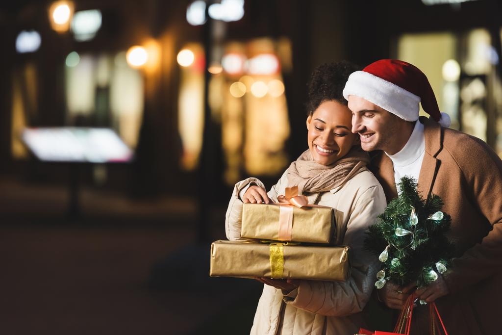 Noel Baba şapkalı mutlu bir adam elinde Noel çantası ve küçük bir çam ağacıyla Afro-Amerikan kız arkadaşının yanında hediye kutularıyla bekliyor. - Fotoğraf, Görsel