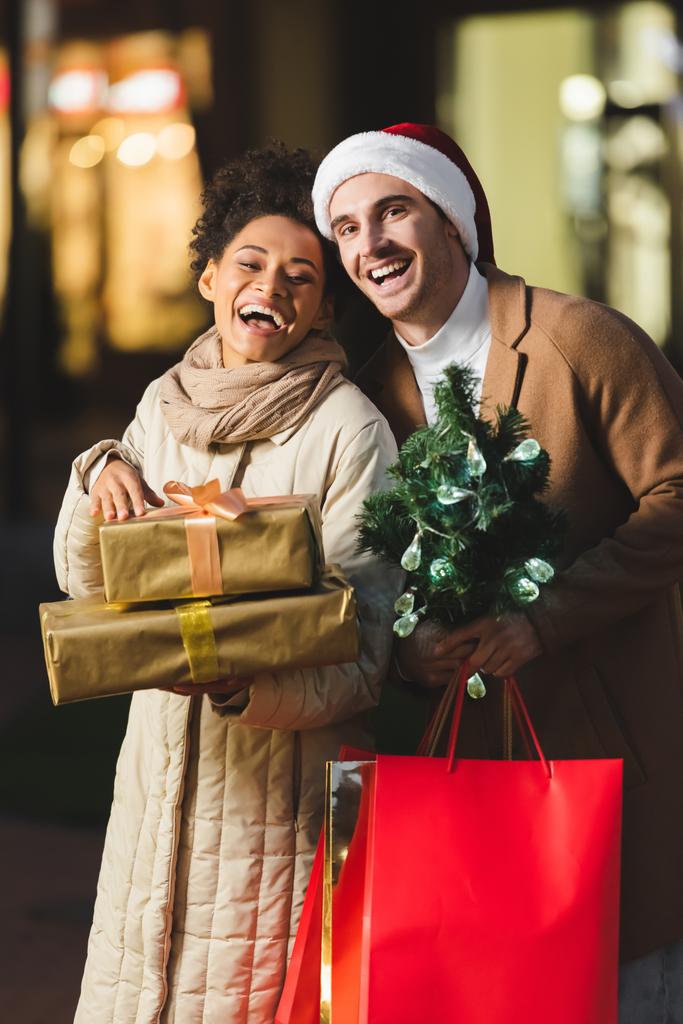 muž v Santa klobouk drží vánoční nákupní tašky a borovice v blízkosti ohromen africké americké přítelkyně s dárkovými krabicemi - Fotografie, Obrázek