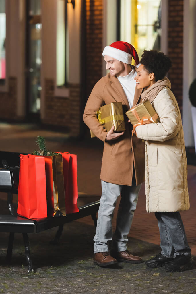 Ευτυχισμένος άντρας με καπέλο Σάντα και αφροαμερικάνικη φιλενάδα να κοιτάει τσάντες για ψώνια ενώ κρατάει Χριστουγεννιάτικα κουτιά δώρων. - Φωτογραφία, εικόνα