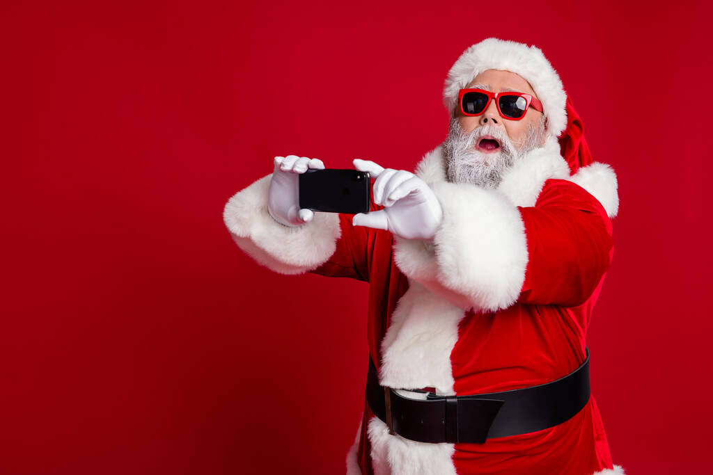 Πορτρέτο της ελκυστική γενειοφόρος έκπληκτος αστείο Σάντα χρησιμοποιώντας τη συσκευή λήψης κάνοντας selfie vlog απομονώνονται σε φωτεινό κόκκινο χρώμα φόντο - Φωτογραφία, εικόνα