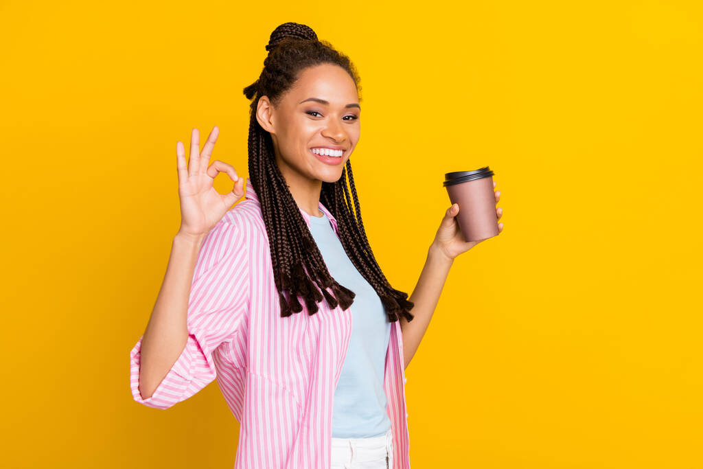Photo of young happy positive dark skin woman make okay sign hold coffee cup wear striped shirt isolated on yellow color background - Photo, Image