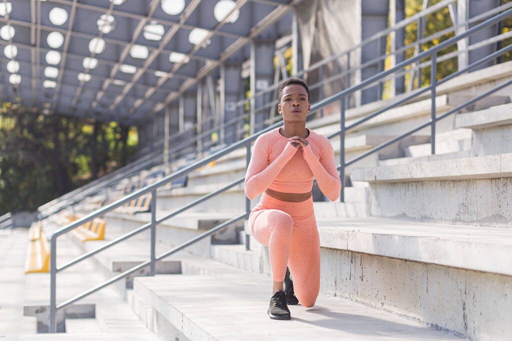 African American woman performs fitness exercises for weight loss, performs squats in a pink suit in the morning in the air near the stadium - Photo, Image