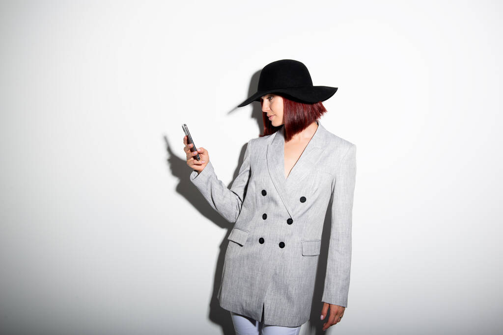 Νεαρή κομψή γυναίκα που χρησιμοποιεί smartphone, εσωτερική πορτρέτο πάνω από λευκό. Μοντέρνο κορίτσι της μόδας που φοράει γκρι μπουφάν και μαύρο καπέλο στέλνει μηνύματα στα κινητά της. - Φωτογραφία, εικόνα
