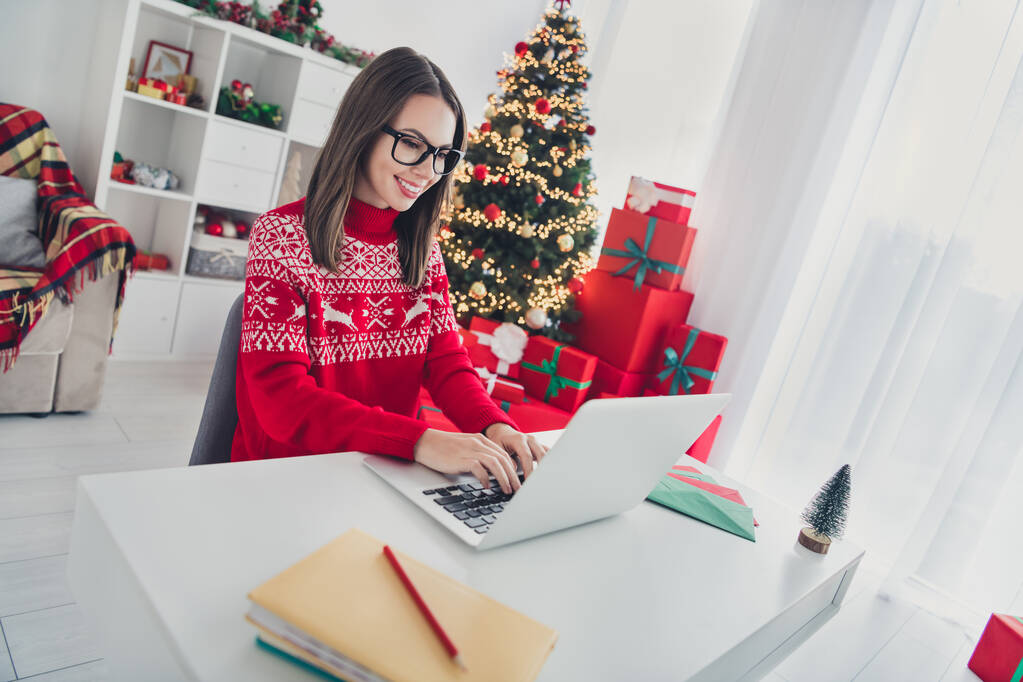 Serbest çalışan bir kadının fotoğrafı çalışma yerinde oturup klavye yazarken Noel Baba şapkalı süveteri giyip kapalı bir evde Noel Baba süveteri giymek. - Fotoğraf, Görsel