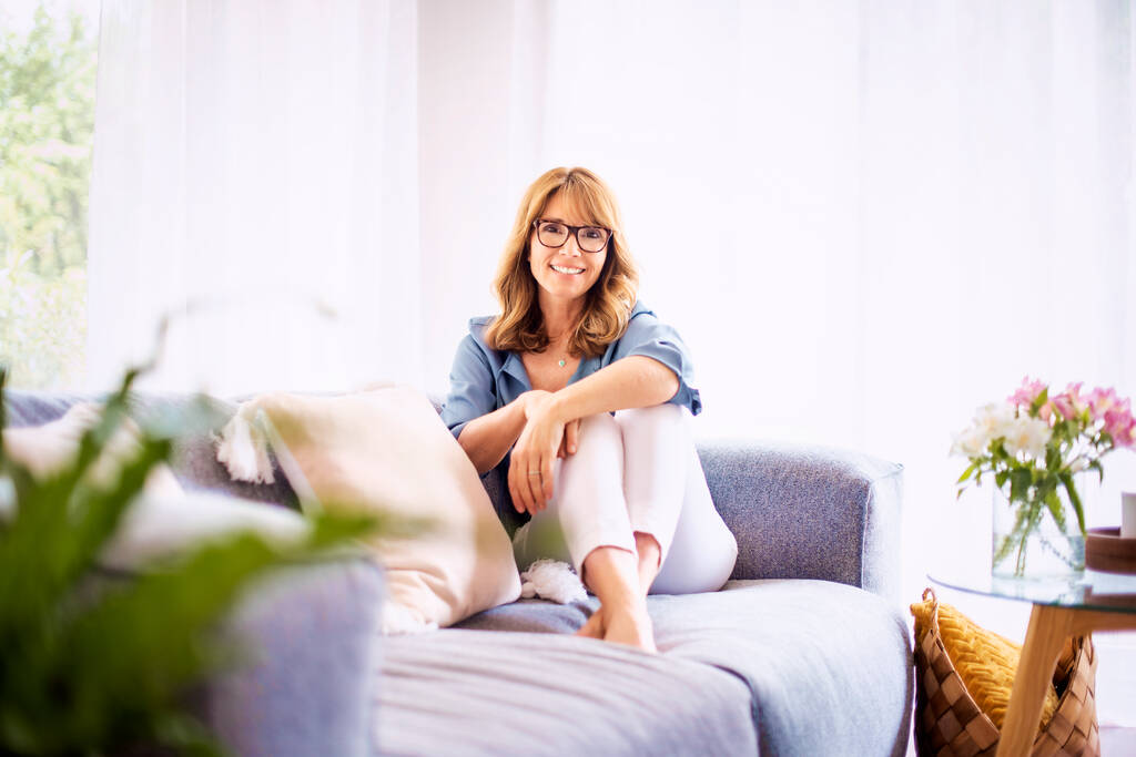 Снимок привлекательной женщины средних лет, смотрящей в камеру и улыбающейся, расслабляясь на диване дома.  - Фото, изображение