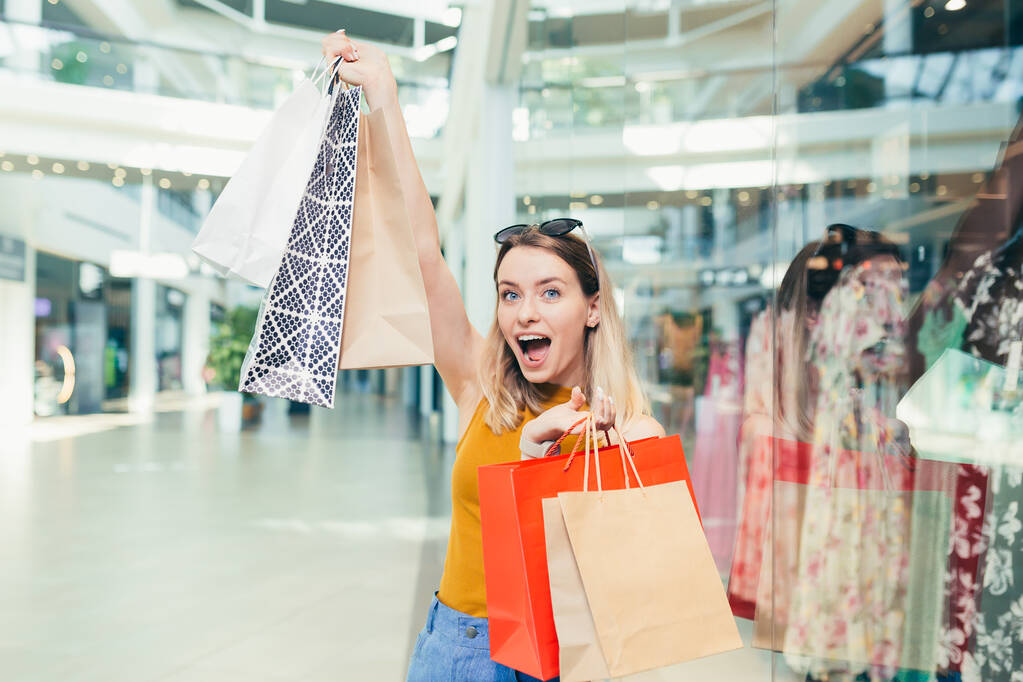 Eine fröhliche junge Shopaholic-Frau hält Papiertüten mit Einkäufen in der Hand und lächelt in die Kamera, die auf einem Einkaufszentrum tanzt. Glückliche Dame, die am schwarzen Freitag Spaß hat, springt freudig vor Freude in die Mall - Foto, Bild
