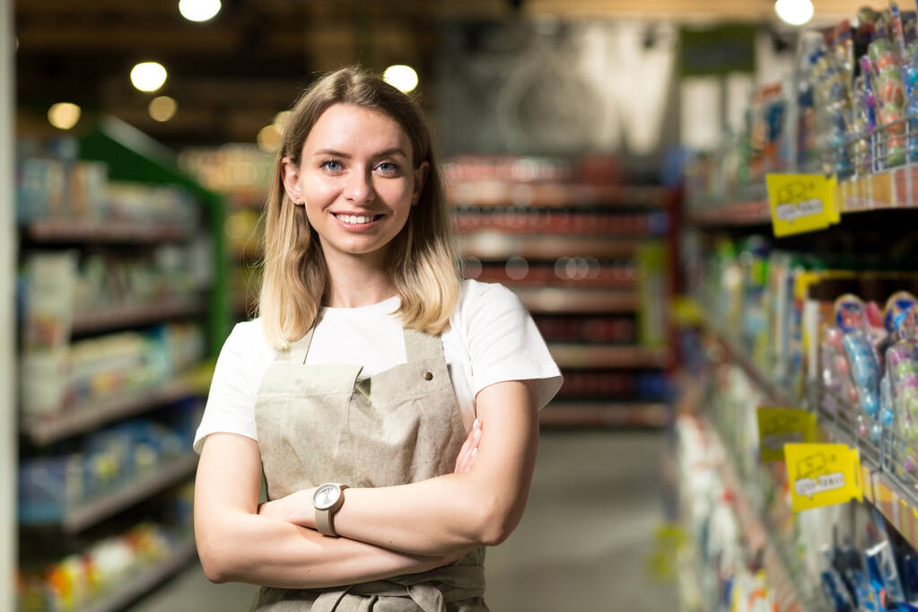 Porträt einer Verkäuferin, einer lächelnden Frau, die im Supermarkt in die Kamera blickt. Angenehm freundliche Verkäuferin, die im Laden zwischen den Reihen steht. Gewerbe und Menschen - Foto, Bild