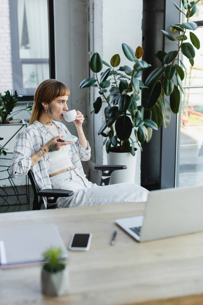 tattooed businesswoman drinking coffee while sitting near work desk and potted plant - Photo, Image