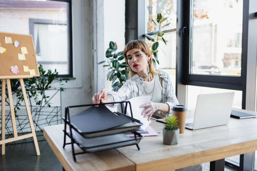 ノートパソコンとコーヒーの近くの紙で仕事をして机の上に行く若い入れ墨のビジネスマンの女性 - 写真・画像