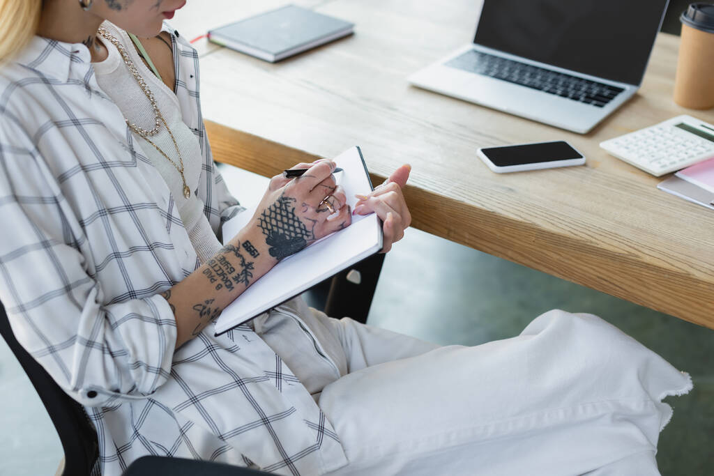 vista recortada de mujer de negocios tatuada escribiendo en cuaderno cerca de gadgets con pantalla en blanco en el escritorio - Foto, Imagen