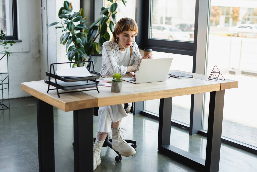 wytatuowana bizneswoman z drinkiem na wynos pracująca w laptopie w nowoczesnym biurze - Zdjęcie, obraz