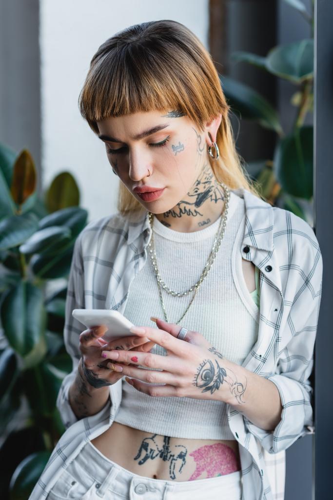 tattooed woman in silver necklaces and piercing messaging on mobile phone in office - Photo, Image