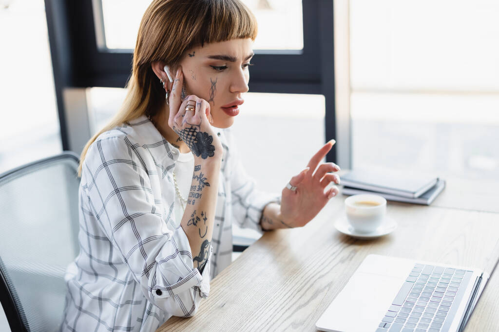 femme tatouée dans l'écouteur assis sur le lieu de travail pendant la vidéoconférence sur ordinateur portable - Photo, image