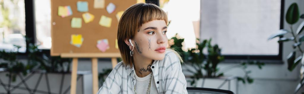 νεαρή γυναίκα με τατουάζ στο ασύρματο ακουστικό κοιτάζει μακριά στο γραφείο, πανό - Φωτογραφία, εικόνα