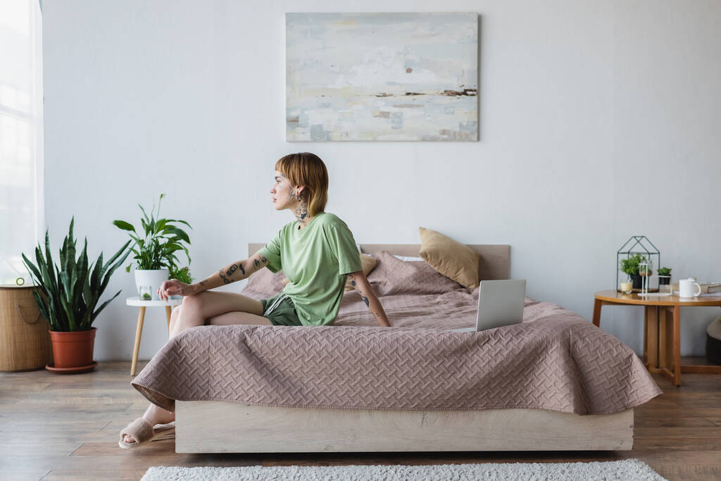 junge tätowierte Frau sitzt auf Bett neben Laptop in modernem Schlafzimmer - Foto, Bild