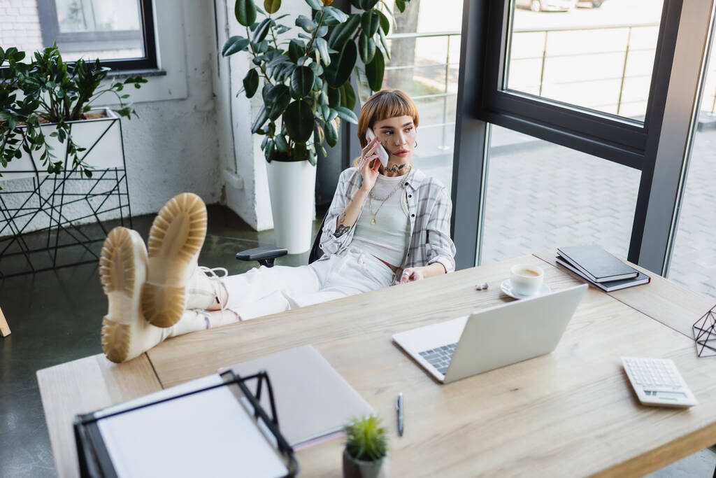 Hochwinkelaufnahme einer tätowierten Frau mit Beinen auf dem Schreibtisch, die mit dem Handy im Büro spricht - Foto, Bild