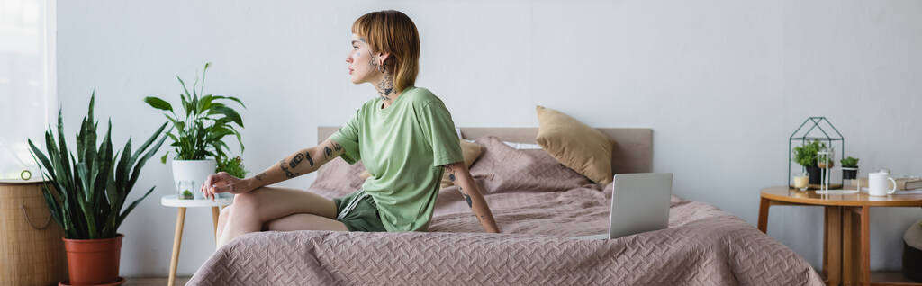 молодая татуированная женщина сидит на кровати возле ноутбука в современной спальне с растениями в горшках, баннер - Фото, изображение