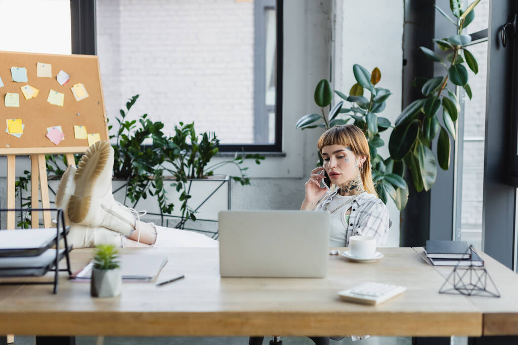 trendy zakenvrouw bellen op mobiele telefoon terwijl zitten met benen op het bureau in de buurt wazig laptop - Foto, afbeelding