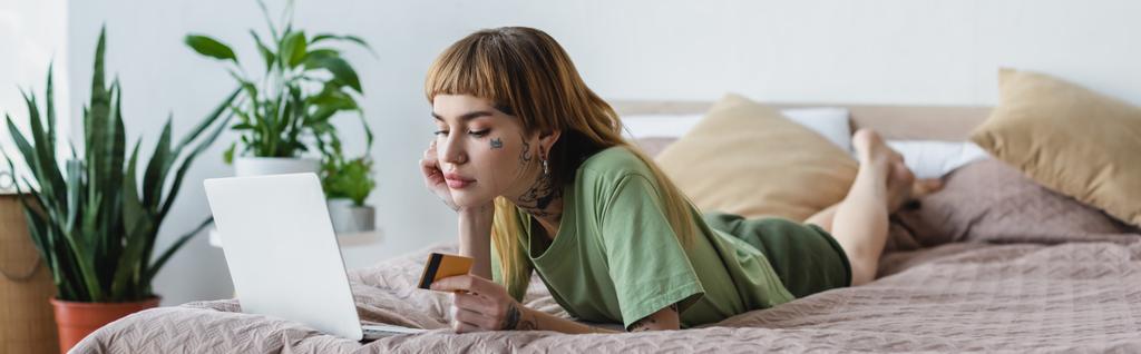 νεαρή γυναίκα με τατουάζ που κρατά πιστωτική κάρτα, ενώ βρίσκεται στο κρεβάτι κοντά στο laptop, πανό - Φωτογραφία, εικόνα