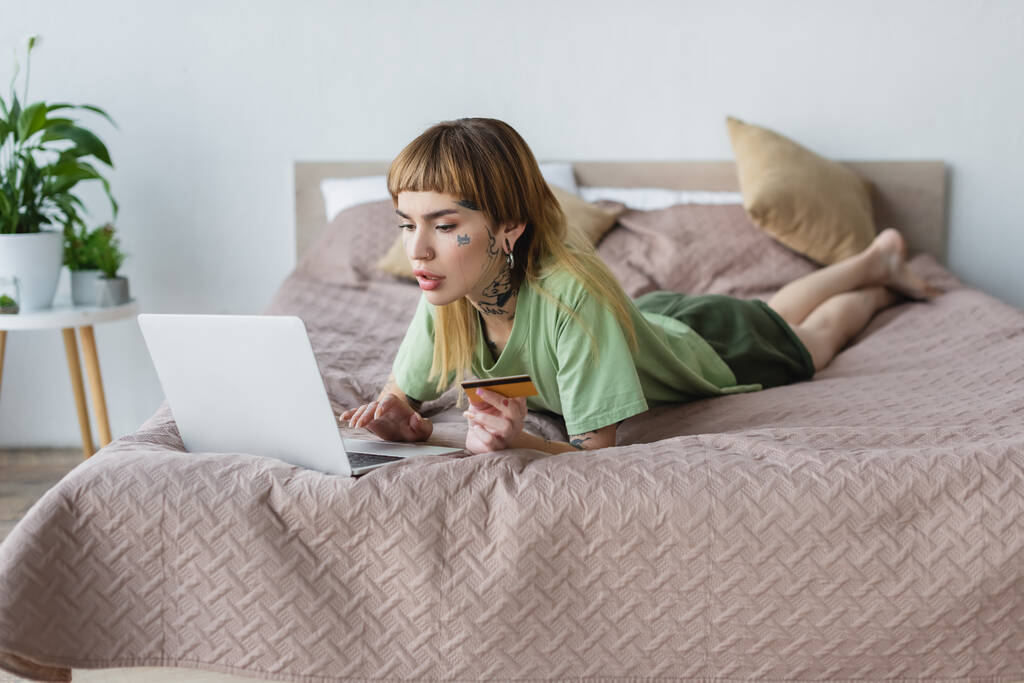 mujer joven con el cuerpo tatuado acostado en la cama con tarjeta de crédito y portátil - Foto, Imagen