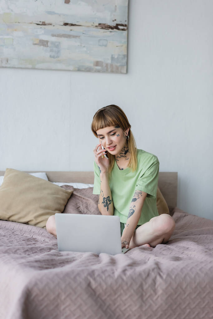 femme tatouée souriante travaillant sur un ordinateur portable et parlant sur un téléphone mobile sur le lit à la maison - Photo, image