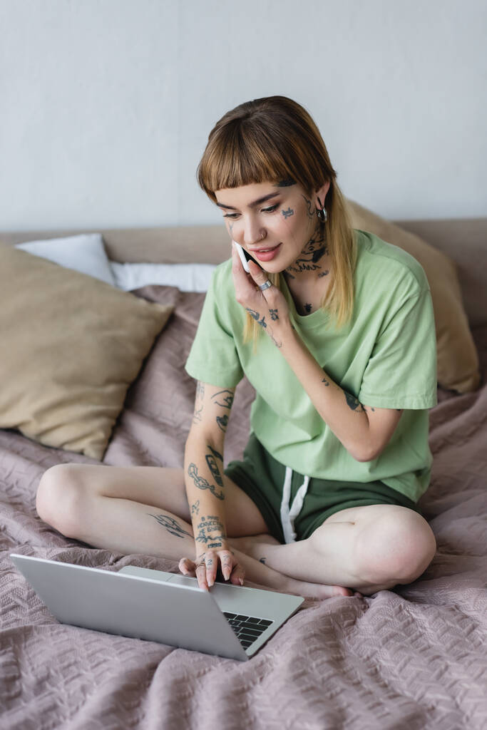 mujer positiva con el cuerpo tatuado escribiendo en el ordenador portátil mientras que llama en el teléfono celular en el dormitorio - Foto, imagen